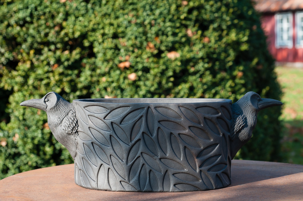 Oval kruka i svart lera med råkhuvuden.